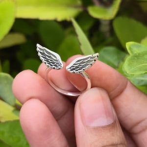 May include: A silver ring featuring an open design with two detailed wing motifs. The wings are textured with feather detailing, creating a delicate and symbolic piece of jewelry. The ring is shown against a backdrop of green foliage.