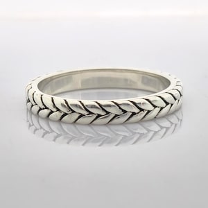 Könnte beinhalten: Ein silberner Ring mit geflochtenem Design. Die Oberfläche des Rings weist ein sich wiederholendes Muster aus ineinander verschlungenen Gliedern auf, wodurch ein strukturiertes Aussehen entsteht. Der Ring wird auf einer reflektierenden Oberfläche präsentiert.