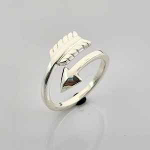 Może przedstawiać: Srebrny pierścionek z motywem strzały. Strzała ma szczegółowe pióra i spiczasty grot. Pierścionek owija się wokół palca, tworząc wyjątkowy i stylowy dodatek.