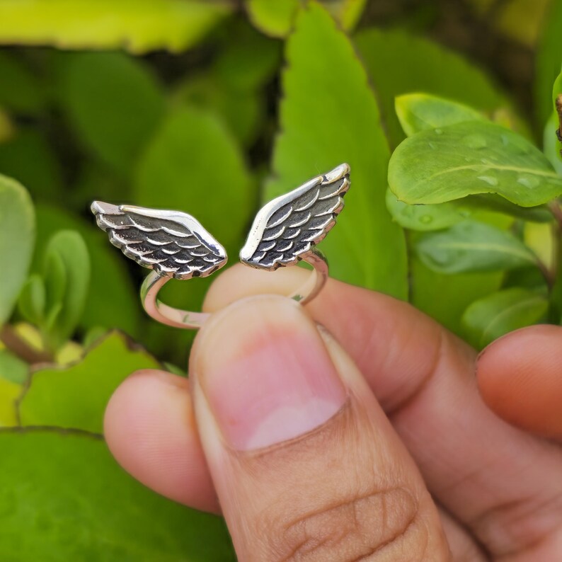 May include: A silver ring featuring two detailed wing designs. The wings are open, creating a unique and symbolic aesthetic. The ring is held against a backdrop of green foliage, highlighting the jewelry's intricate details.