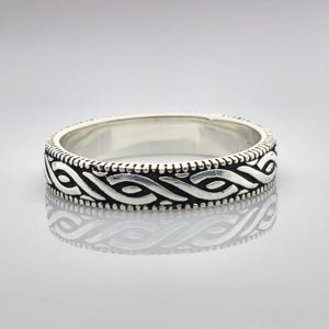 May include: A silver ring with a detailed, interwoven pattern around the band. The design features a series of interconnected loops and lines, creating a textured surface. The ring's surface reflects light, highlighting the intricate craftsmanship.