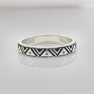 Puede incluir: Un anillo plateado con un diseño geométrico. La banda presenta un patrón repetitivo de triángulos negros y estrellas. El anillo es reflectante y está sobre una superficie clara. El anillo mide aproximadamente 0,5 cm de ancho.