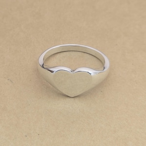 Op de afbeelding: Een gepolijste zilveren ring met een hartvormige voorkant. De ring heeft een gladde, afgeronde band en een effen hartontwerp. De ring wordt getoond tegen een neutrale, bruinachtige achtergrond.