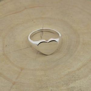 Può includere: Un anello d'argento lucido con una forma a cuore. La superficie dell'anello è liscia e riflettente. Il cuore è pieno e centrato sulla fascia. L'anello è appoggiato su una superficie di legno marrone chiaro.
