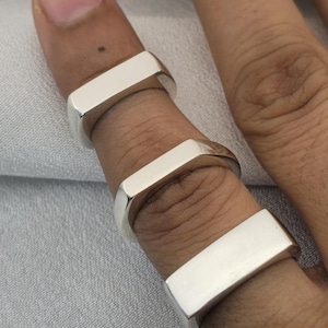 Op de afbeelding: Drie zilveren ringen met een rechthoekig ontwerp worden aan vingers gedragen. De ringen hebben een gepolijst, reflecterend oppervlak. De vingers bevinden zich tegen een lichtgrijze achtergrond.