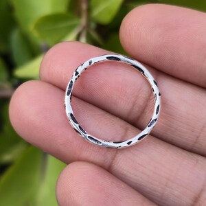 Op de afbeelding: Een zilveren ring met een getextureerd oppervlak en donkere accenten. De ring is cirkelvormig en lijkt handgemaakt. De ring wordt in een hand gehouden tegen een vage groene achtergrond.