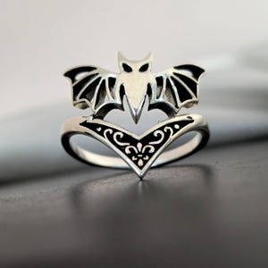 May include: A silver ring featuring a bat design. The bat has outstretched wings and is positioned above a decorative, heart-shaped base with black filigree. The ring is a gothic-inspired piece of jewelry.