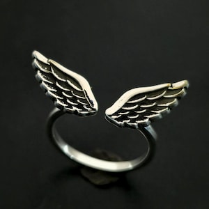 May include: A silver-colored ring featuring an open design with two detailed wing shapes. The wings are positioned to appear as if they are wrapping around a finger. The ring is set against a dark background, highlighting the metallic sheen.