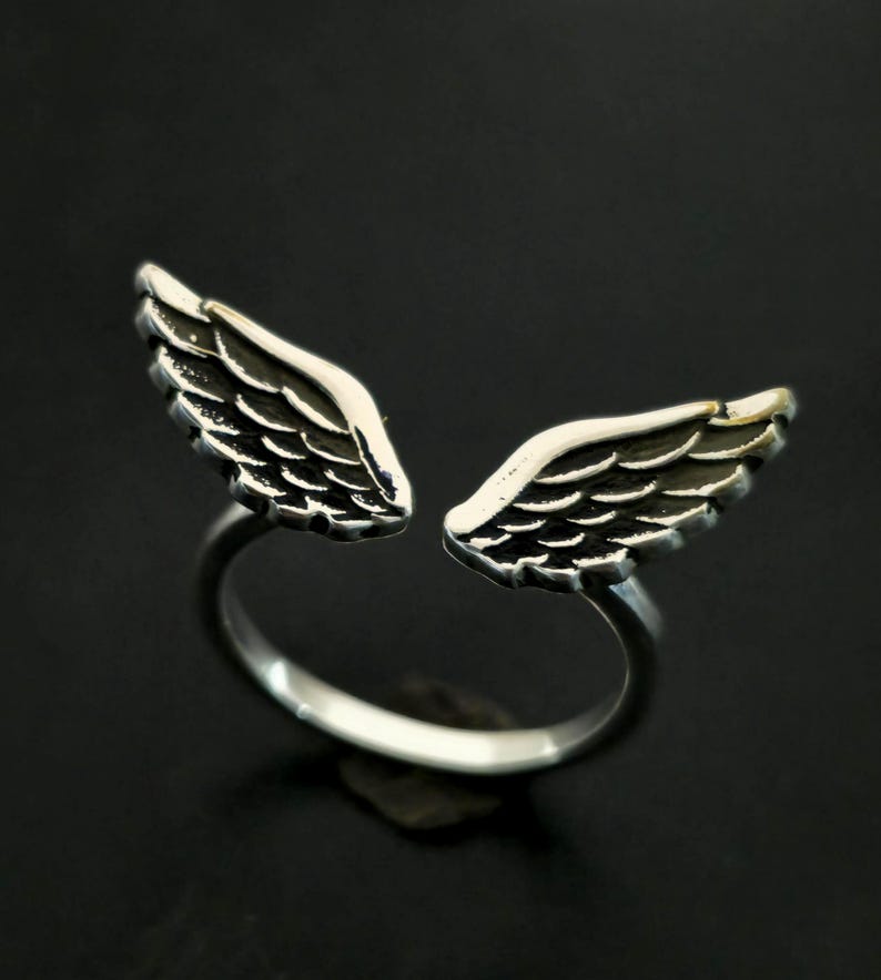 May include: A silver-colored ring featuring an open design with two detailed wing shapes. The wings are positioned to appear as if they are wrapping around a finger. The ring is set against a dark background, highlighting the metallic sheen.