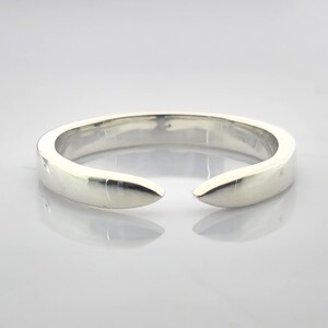 Op de afbeelding: Een zilverkleurige manchetarmband met open uiteinden. De armband heeft een glad, afgerond ontwerp met een opening aan de voorkant. Het metaal reflecteert licht, waardoor highlights en schaduwen ontstaan. Het is een eenvoudig, modern sieraad.