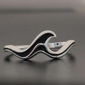 Puede incluir: Un anillo de plata con diseño de ola. El anillo presenta una banda de plata pulida con un patrón de ola de esmalte negro. Las crestas y valles de las olas están claramente definidos, creando una joya elegante y única.