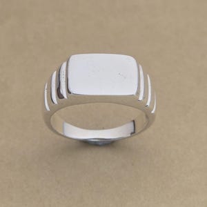 Op de afbeelding: Een zilveren zegelring met een rechthoekige voorkant en een gepolijste afwerking. De ring heeft een klassiek ontwerp met drie verticale groeven aan weerszijden van de voorkant. De ring wordt op een neutrale achtergrond getoond.