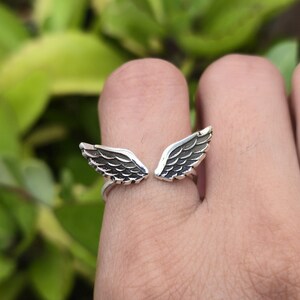May include: A silver ring featuring an open wing design. The wings are detailed with etched feather patterns. The ring is worn on a finger, with a blurred green background.