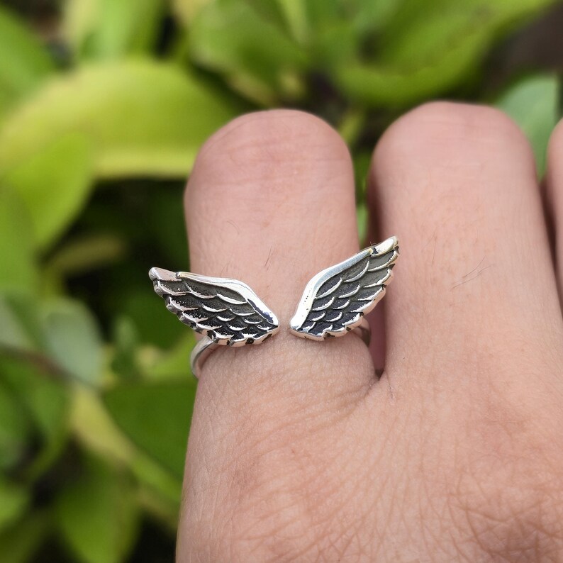May include: A silver ring featuring an open wing design. The wings are detailed with etched feather patterns. The ring is worn on a finger, with a blurred green background.