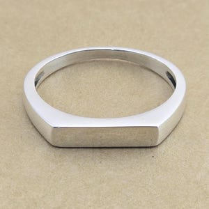 Puede incluir: Un anillo de plata con una cara rectangular y una banda redondeada. El anillo tiene un acabado pulido, que refleja la luz. El diseño es simple y moderno, adecuado para el uso diario. Es una pieza de joyería.