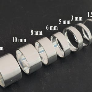 Puede incluir: Una colección de anillos de plata de diferentes anchos, que van desde 1,5 mm hasta 13 mm. Los anillos se muestran sobre una superficie oscura, mostrando sus superficies pulidas y reflectantes. Los tamaños están etiquetados.