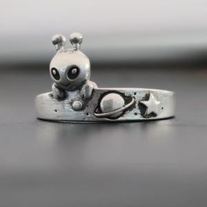 Puede incluir: Un anillo de plata con una pequeña figura de alienígena sonriente. La banda del anillo tiene un diseño de planeta y estrella. El alienígena tiene ojos grandes y dos antenas. El anillo probablemente está hecho de plata de ley.