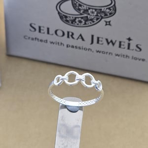 May include: A silver ring with three linked circles across the top. The band has a crisscross pattern. The background includes the text "SELORA JEWELS" and "Crafted with passion, worn with love."