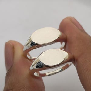 Puede incluir: Dos anillos de sello plateados con caras ovaladas se sostienen sobre un fondo claro. Los anillos tienen una superficie pulida y reflectante. Las caras ovaladas son lisas y sin adornos, reflejando la luz.
