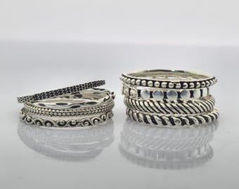 May include: Two stacks of silver rings are displayed, each with a unique design. One stack includes a ring with small black stones. The other features rings with beaded, twisted, and patterned details. The rings are presented on a reflective surface.
