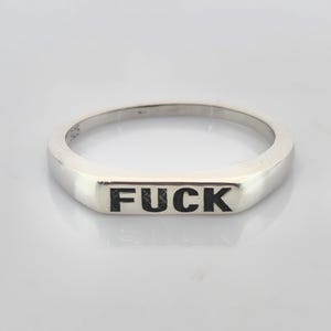 Può includere: Un anello d'argento con la parola "FUCK" in grassetto nero. L'anello ha una faccia rettangolare e una fascia lucida. L'anello è probabilmente in argento sterling, come indicato dal timbro "925".
