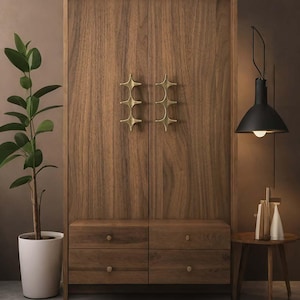 May include: A wooden cabinet with star-shaped brass handles. The cabinet has six drawers and is next to a potted plant and a small side table with a black lamp. The cabinet is made of dark wood.