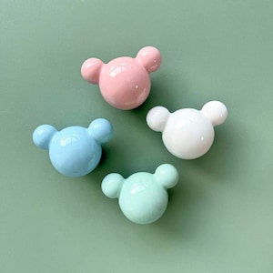 Könnte beinhalten: Vier Keramik-Schubladengriffe in Form eines Bärenkopfes. Die Griffe sind in Pastellfarben: rosa, weiß, hellblau und hellgrün.