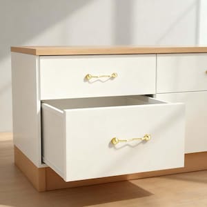 Op de afbeelding: Een witte dressoir met een licht houten blad en voet. Eén lade is open en onthult de binnenkant. De laden hebben goudkleurige, gebogen handgrepen. De dressoir heeft een moderne stijl en lijkt nieuw.