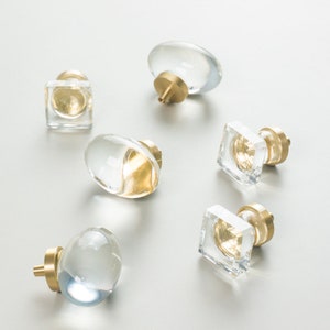 May include: Assortment of six clear glass cabinet knobs with brass accents. The knobs are in various shapes, including round, oval, and square.