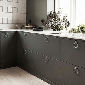 May include: Dark gray kitchen cabinets with silver ring pulls. The cabinets are set against a white countertop and a white tiled backsplash. A stainless steel faucet and sink are visible near a window with greenery.