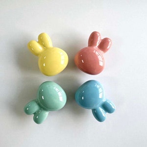 May include: Four ceramic bunny figurines in pastel colours: yellow, pink, light blue and teal. The bunnies are all facing the same direction with their ears up.