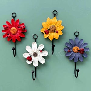 Może przedstawiać: Cztery dekoracyjne haczyki ścienne w kształcie kwiatów. Haczyki mają kształt czerwonej stokrotki, żółtej słonecznika, białej stokrotki i niebieskiej stokrotki. Każdy kwiat ma czarny haczyk i jest ozdobiony biedronkami.