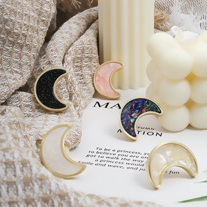 May include: Five gold crescent moon shaped brooches with different coloured inlays. The inlays are black with glitter, pink, blue and green marbled, white, and white with gold flecks. The brooches are on a white surface with text that reads "To be a princess, you to believe you are on got to walk the way a princess would smile and wave, and ju: fun"