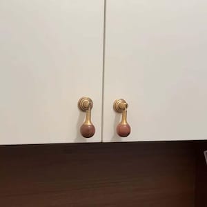 May include: Two gold cabinet knobs with wooden ball finials. The knobs are mounted on a white cabinet door.