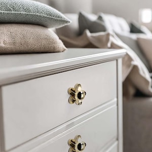 Puede incluir: Primer plano de una cómoda blanca con dos cajones, cada uno adornado con un pomo dorado en forma de flor. La cómoda está rematada con dos almohadas texturizadas en tonos grises y beige. El fondo muestra una cama con almohadas.