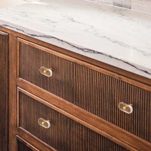 May include: A wooden cabinet with three drawers, featuring a vertical grooved design. The cabinet is fitted with clear, oval-shaped knobs with gold accents. A white marble countertop with grey veining is positioned above the cabinet.