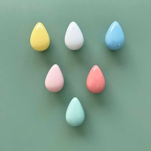 Peut inclure: Six crochets en céramique en forme de goutte d'eau dans différentes couleurs pastel : jaune, blanc, bleu clair, rose, corail et vert clair. Les crochets sont disposés en forme de triangle sur un fond vert.