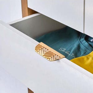 May include: A white drawer with a gold-colored leaf-shaped handle. The drawer is open, revealing a teal shirt and a yellow shirt inside. The handle has a cut-out design.