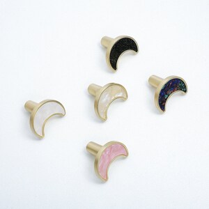 Może przedstawiać: Pięć półksiężycowych gałek do szafek z mosiężną podstawą. Gałki mają różne kolorowe wkładki: czarne, białe, różowe, kremowe i niebieskie.