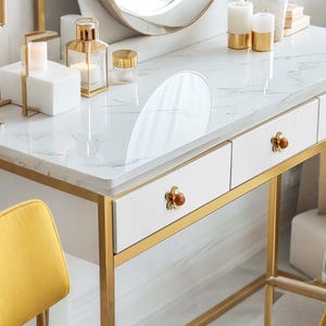 May include: A white vanity table with a marble top and gold-colored metal frame. The table has two drawers with flower-shaped knobs. Decorative candles, a mirror, and a yellow chair are also visible.
