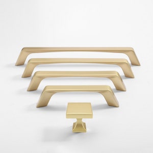 May include: Five gold cabinet pulls in different sizes, arranged in descending order from left to right. A smaller gold cabinet knob is in the centre of the image.