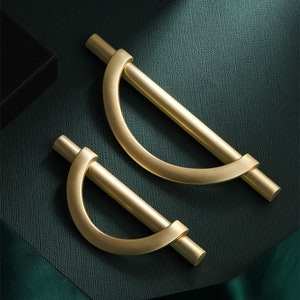 May include: Two gold-toned cabinet pulls with a half-circle design. The pulls are made of metal and have a brushed finish.