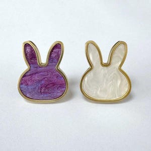May include: A pair of bunny-shaped earrings. One earring has a purple and white marbled design with gold-coloured trim. The other has a white marbled design with gold-coloured trim. The earrings are on a white background.