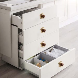Op de afbeelding: Witte kast met vier lades, elk met een bloemvormige goudkleurige knop met een bruin centrum. De onderste lade is open en toont babyartikelen. De kast maakt deel uit van een keuken of kinderkamer.