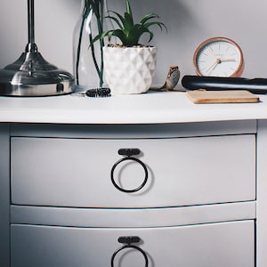 May include: Two white drawers with black ring-shaped pulls. The top drawer is open and the bottom drawer is closed. The drawers are on a white bedside table with a lamp, a vase of flowers, a potted plant, a watch, a hair straightener, and a wooden coaster.