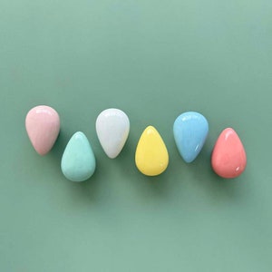 Może przedstawiać: Siedem ceramicznych gałek w kształcie łezki w pastelowych kolorach: różowym, miętowym, białym, żółtym, jasnoniebieskim i koralowym. Gałki ułożone są na jednolitym zielonym tle, stanowiąc kolorowy element dekoracyjny.