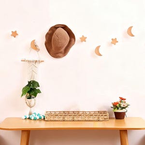 Puede incluir: Una decoración de pared con estrellas y lunas crecientes de madera. Un sombrero de sol tejido cuelga de la pared, junto con un colgador de plantas de macramé con una planta verde. Una cesta tejida y una planta en maceta con flores están sobre una mesa de madera.