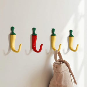 Może przedstawiać: Cztery haczyki ścienne w kształcie papryki chili. Trzy są żółte z zielonymi łodygami, a jeden jest czerwony z zieloną łodygą. Beżowa torba wisi na jednym z żółtych haczyków. Haczyki są zamontowane na białej ścianie.