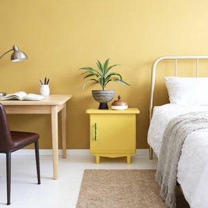 May include: A bedroom interior featuring a yellow bedside cabinet, a wooden desk, and a bed with white bedding. A plant in a pot sits on the cabinet. The room has a yellow wall and a brown rug. A silver lamp is on the desk.