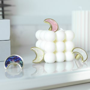 Puede incluir: Cuatro tiradores decorativos para cajones en forma de media luna con acabado dorado e incrustaciones iridiscentes. Los tiradores se muestran en una vela blanca con textura de burbujas.